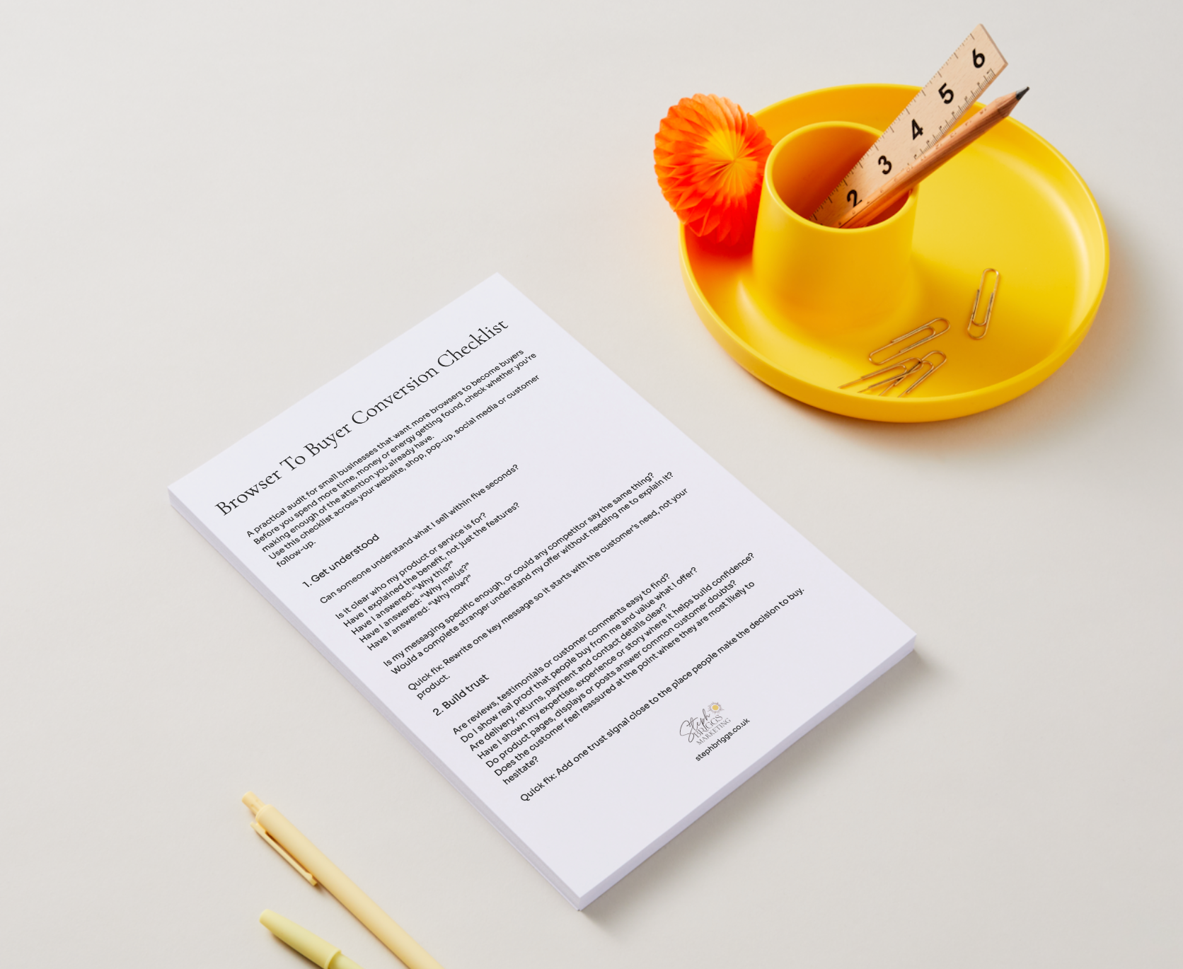 A printed checklist sheet titled 'Browser To Buyer Conversion Checklist' lies on a light neutral background, styled with a yellow cup, orange pom-pom, paperclips, and pens.