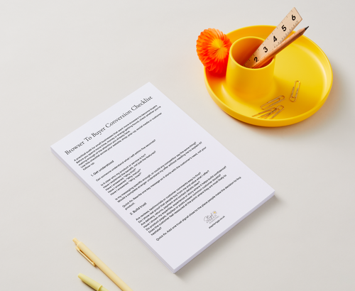 A printed checklist sheet titled 'Browser To Buyer Conversion Checklist' lies on a light neutral background, styled with a yellow cup, orange pom-pom, paperclips, and pens.
