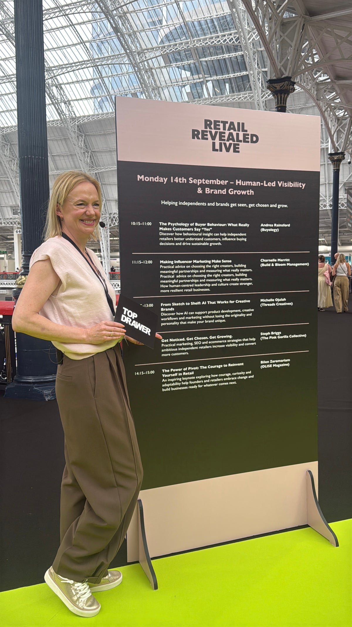A woman stands smiling next to a large vertical event board reading 'RETAIL REVEALED LIVE' at an indoor exhibition hall. She is wearing a light pink sleeveless top, brown wide-leg trousers, and gold trainers, holding a black tote card with 'TOP DRAWER' printed on it.